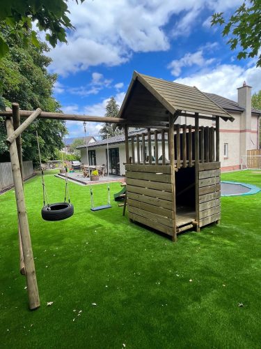 Wooden climbing frame with swing set in the side on astroturf with a new house in the background. Blue skies with some white clouds. Fence and large trees.
